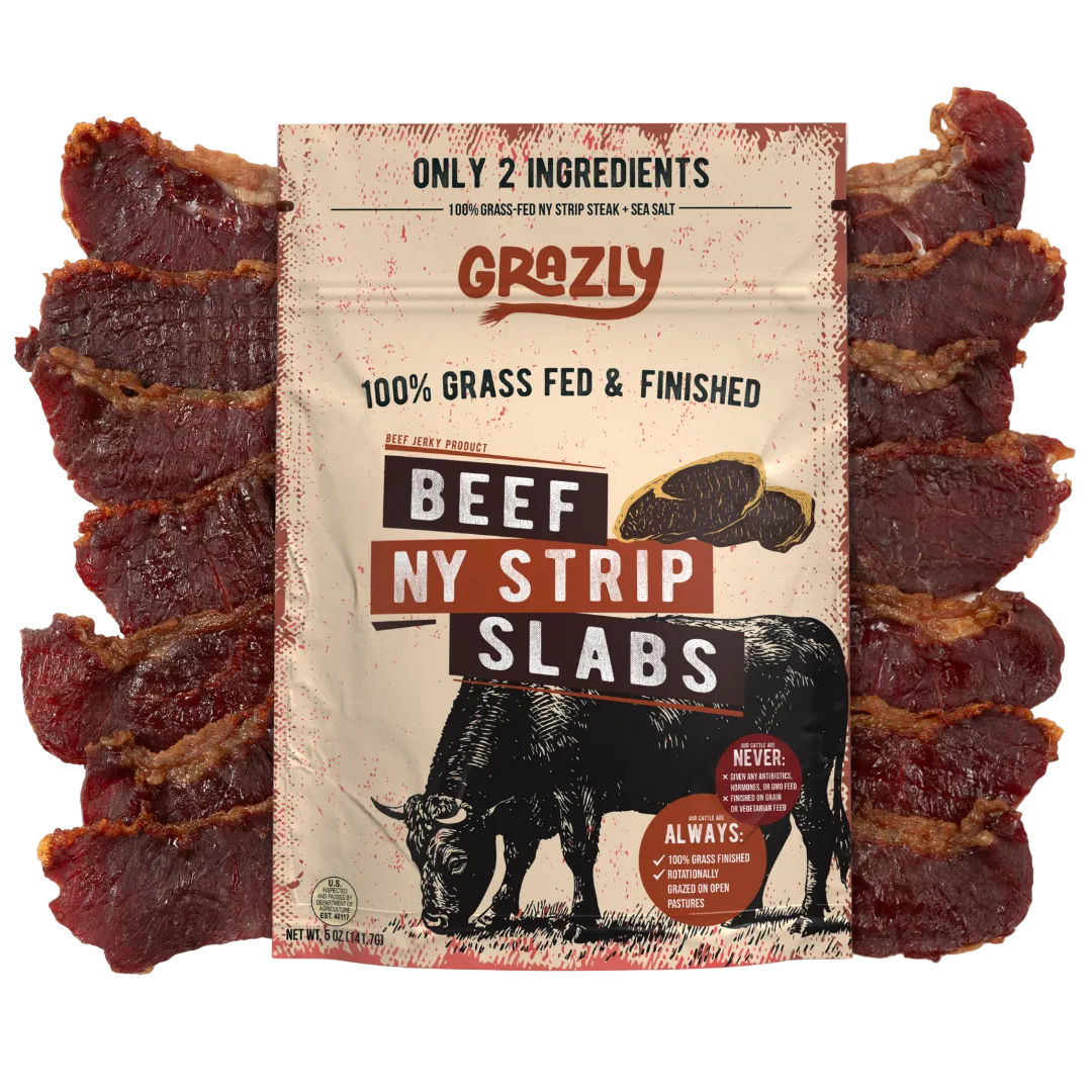A package of Grazly Beef Ny Strip Slabs surrounded by several slices of the marbled beef jerky on a plain white background.